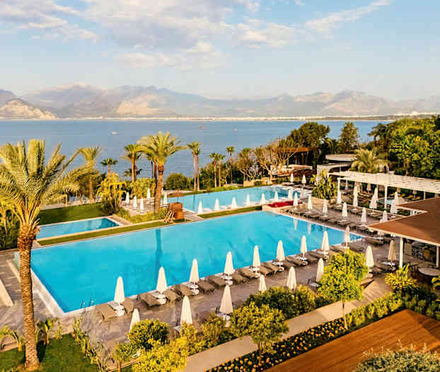 Billede af en stor pool omgivet af liggestole med parasoller, grønt områder og udsigt over havet og bjergene i baggrunden. (AI-genereret tekst)