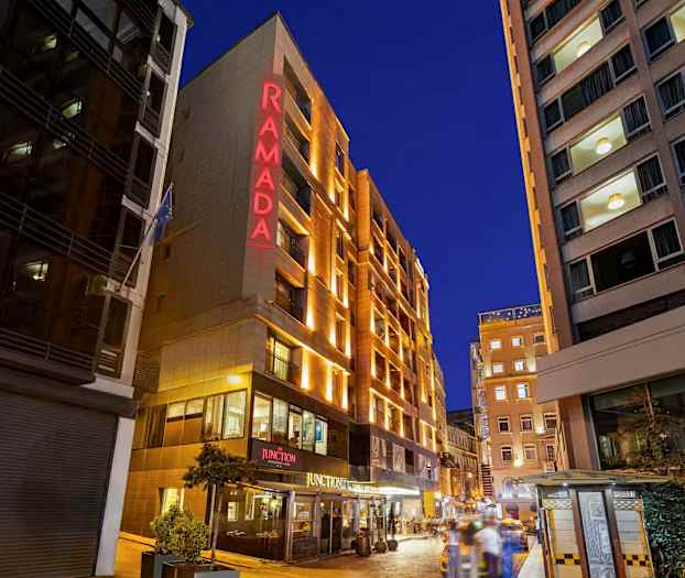 Billedet viser facaden af et hotel med teksten "RAMADA" oplyst i rødt, omgivet af andre bygninger og en aftenoplyst bymiljø. (AI-genereret tekst)