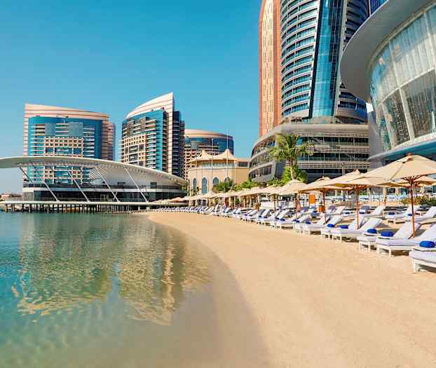 En strand med liggestole og parasoller foran moderne højhuse ved havet. (AI-genereret tekst)