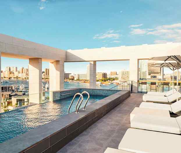 En tagterrasse med pool tilbyder udsigt over en by og en havn, omgivet af solvogne og parasoller. (AI-genereret tekst)