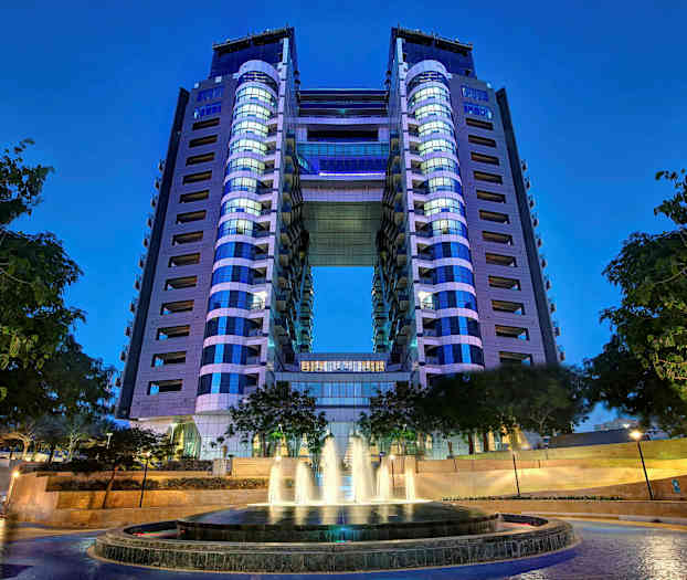 En moderne bygning med to tårne forbundet af en bro. I forgrunden er en springvand omgivet af træer. (AI-genereret tekst)