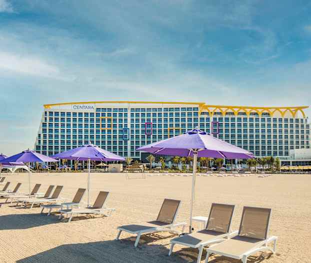 Billedet viser en sandstrand med liggestole og lilla parasoller. I baggrunden ses en stor bygning med flere etager. (AI-genereret tekst)