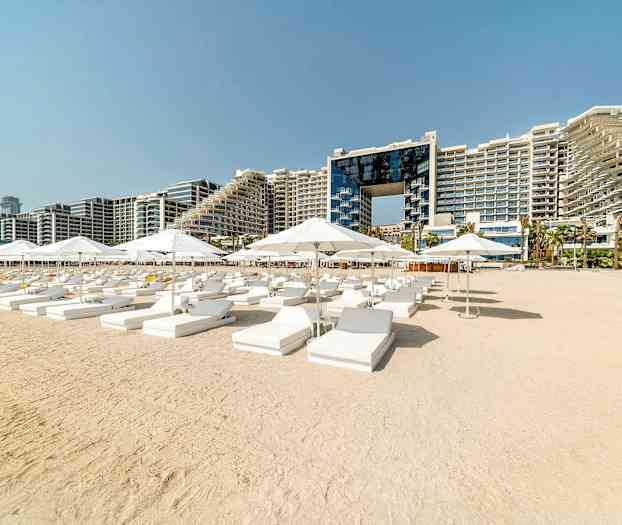 Solstole og parasoller på en sandstrand med et hotelkompleks i baggrunden. (AI-genereret tekst)