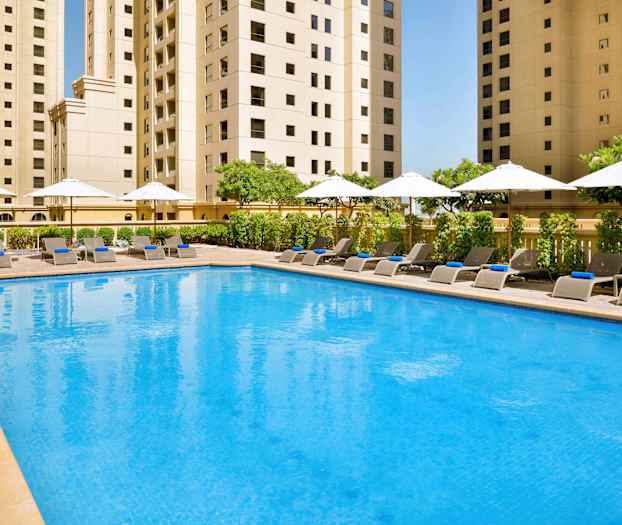 En pool med liggestole og parasoller omgivet af høje bygninger. (AI-genereret tekst)