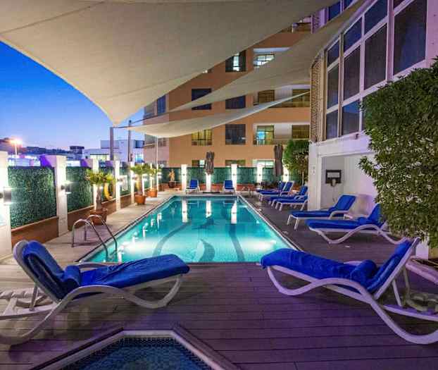 En oplyst swimmingpool med omkringliggende liggestole og planter, under en beskyttende presenning om aftenen. (AI-genereret tekst)