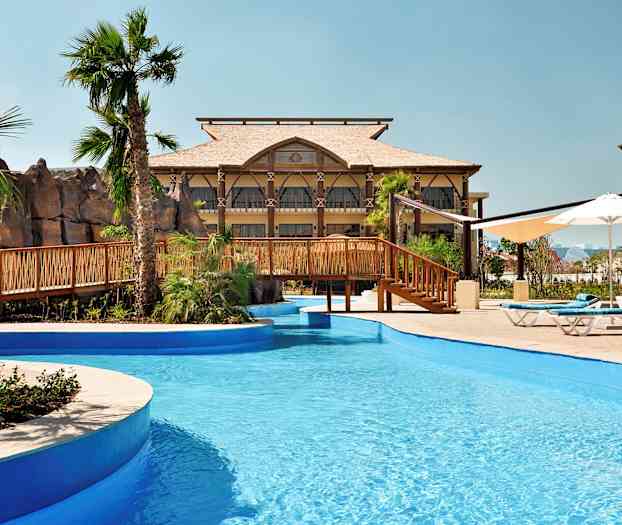 En pool med blåt vand og en træbro, omgivet af palmer og bygninger. (AI-genereret tekst)