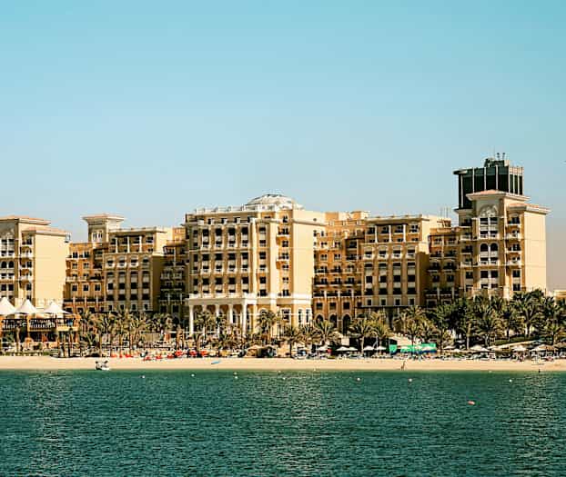 En stor hotelbygning ved en strand med palmer foran, en lys himmel i baggrunden og havet i forgrunden. (AI-genereret tekst)