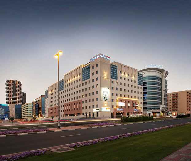 Bymiljø i skumringen med lejlighedsbygninger og moderne hotelbygninger. En vej med biltrafik og blomster langs fortovet. (AI-genereret tekst)