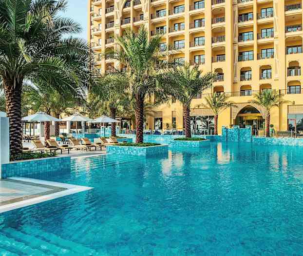 Hotelpool med klart vand, omgivet af palmer og liggestole. (AI-genereret tekst)