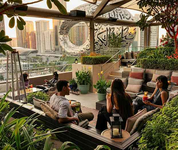 En udendørs lounge med siddepladser omgivet af grønne planter, med en udsigt over byen og en moderne bygning i baggrunden. (AI-genereret tekst)