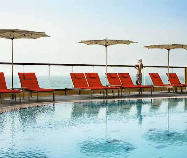 En udendørs pool med røde liggestole og hvide parasoller ved siden af, med havudsigt i baggrunden. (AI-genereret tekst)