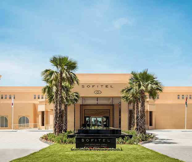 Forsiden af Sofitel-hotellet med palmer og flag. (AI-genereret tekst)