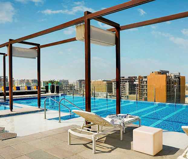 En udendørs pool med udsigt over en by. Liggestole, borde og dekorationer er placeret omkring poolen. (AI-genereret tekst)