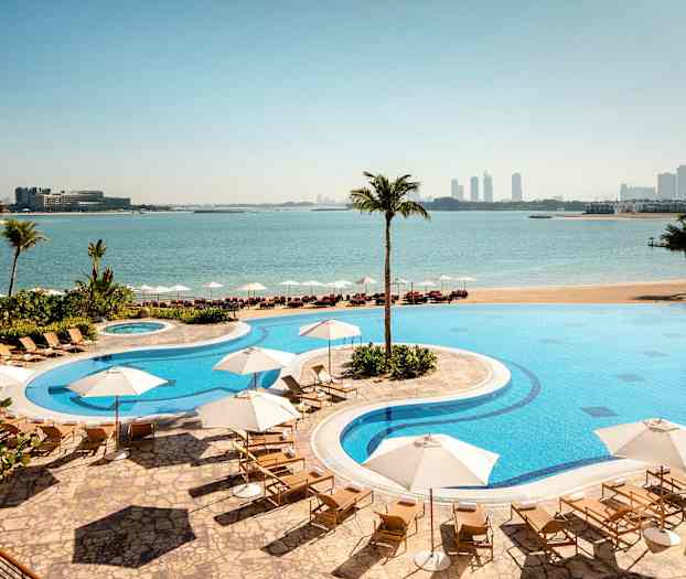 En hotelpool med liggestole og parasoller beliggende ved en strand med udsigt over havet og en by i baggrunden. (AI-genereret tekst)