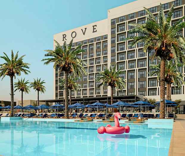 En bygning mærket 'ROVE' bag en pool med palmer og liggestole omkring samt en rosa flamingo oppustelig legetøj i vandet. (AI-genereret tekst)