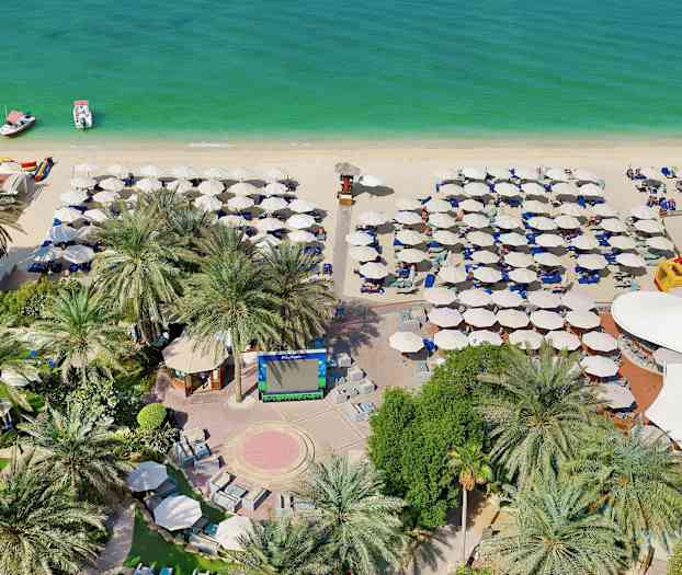 Billede af en strand med parasoller, liggestole, to både på vandet samt palmer og beplantning i forgrunden. (AI-genereret tekst)