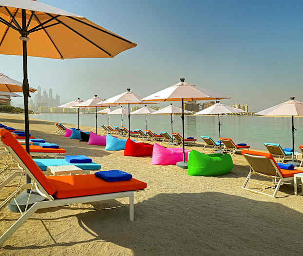 En sandstrand med liggestole med farverige puder og parasoller ved en rolig vandflade. (AI-genereret tekst)