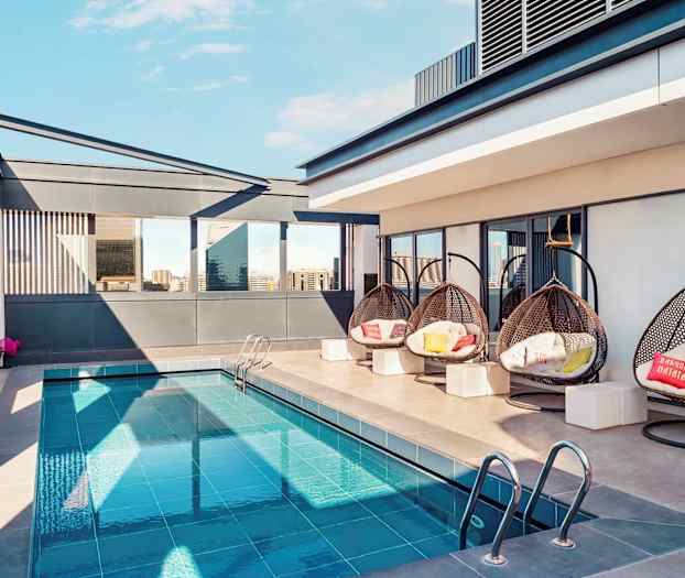 En tagterrasse med en lille pool, hængende flettede stole og moderne omgivelser. Bygninger ses i baggrunden. (AI-genereret tekst)