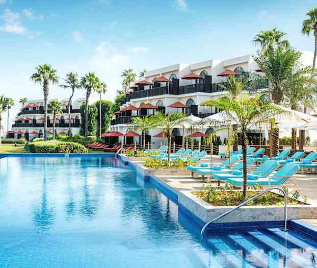 Et hotelområde med flere etager, palmer, en swimmingpool og liggestole under parasoller. (AI-genereret tekst)