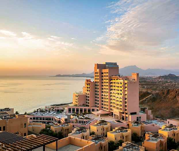 Hotelbygning tæt på havet ved solnedgang, omgivet af mindre bygninger og bjerge i baggrunden. (AI-genereret tekst)