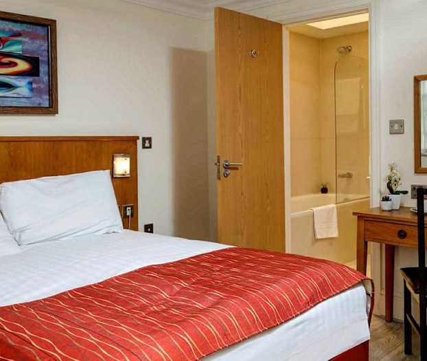 Hotelværelse med seng, skrivebord, spejl og udsigt til et badeværelse. (AI-genereret tekst)