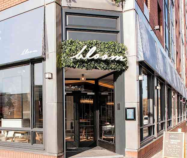 Indgangen til restauranten Bloom med store vinduer og et skilt med navnet omkranset af grønt. (AI-genereret tekst)