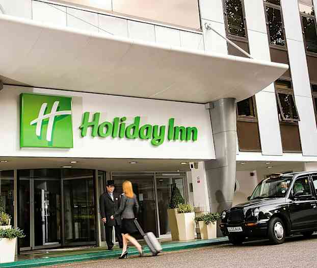 Indgangen til Holiday Inn med en taxa parkeret udenfor og en person, der trækker en kuffert. (AI-genereret tekst)