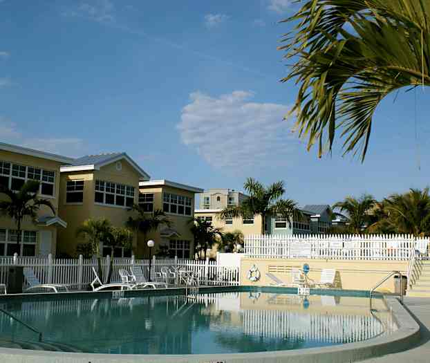 En pool omgivet af liggestole, parasoller og huse, med tropiske palmer i baggrunden. (AI-genereret tekst)