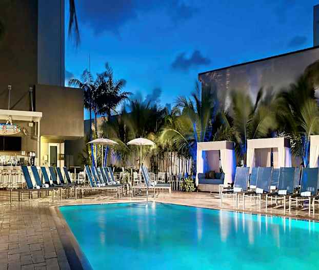 En udendørs pool med liggestole og cabanas omkring samt en bar i baggrunden med palmer rundt om. (AI-genereret tekst)