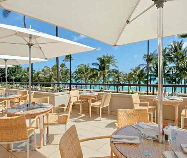 En udendørsterrasse med borde og stole under parasoller, med havudsigt og palmer i baggrunden. (AI-genereret tekst)