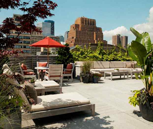 En rummelig tagterrasse med udendørsmøbler, parasol og planter. I baggrunden ses bygninger og en klar himmel. (AI-genereret tekst)