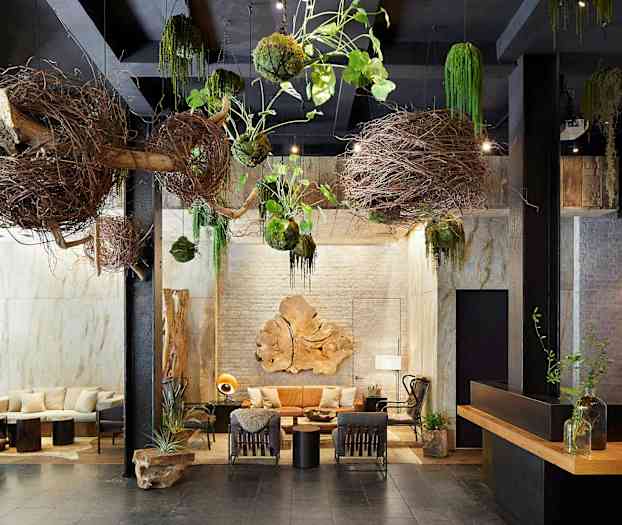 En indendørs interiør med naturlige dekorationer, såsom grene og planter hængende fra loftet. Der er siddemøbler og borde med dekorationer i midten. (AI-genereret tekst)