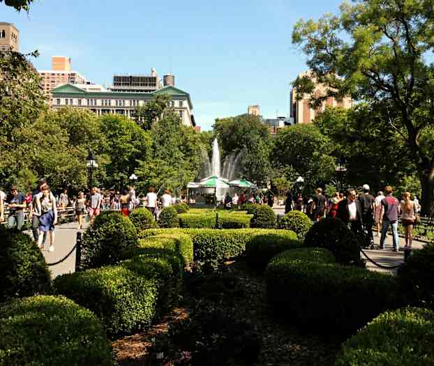 En park med buske, træer og en springvand i baggrunden, hvor folk går og opholder sig. (AI-genereret tekst)
