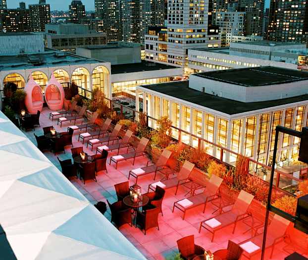 En tagterrasse med solsenge og belysning om aftenen, omgivet af bygninger. (AI-genereret tekst)