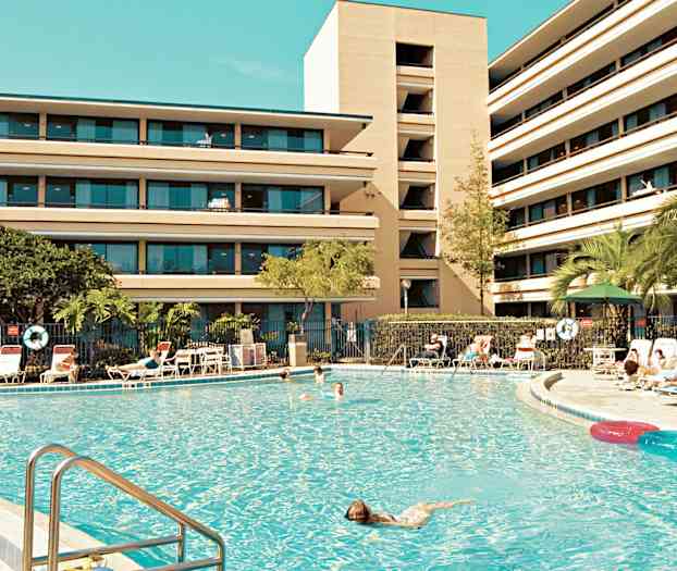 En stor swimmingpool omgivet af liggestole og en bygning med flere etager i baggrunden. (AI-genereret tekst)
