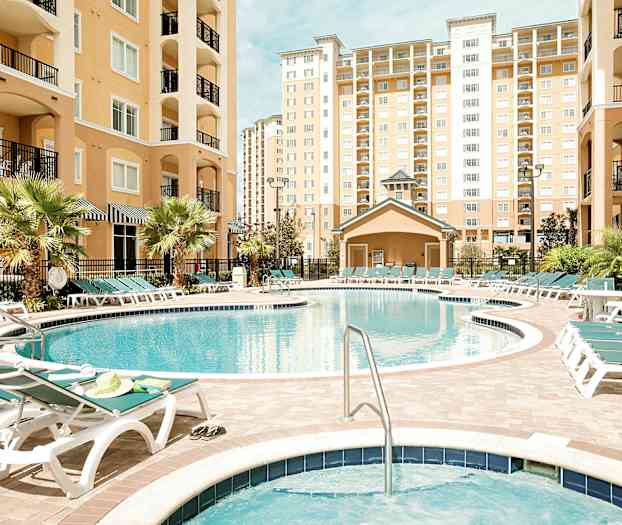 Billede af en udendørs swimmingpool omgivet af liggestole og omkranset af bygninger med flere etager. (AI-genereret tekst)