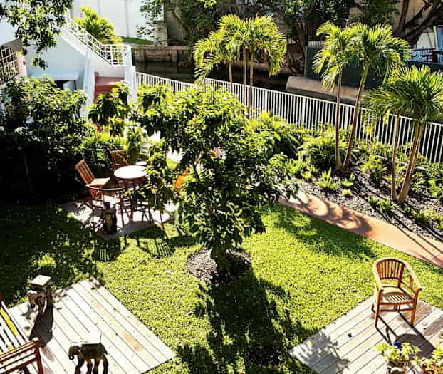 Udsigt over en grøn have med flere træer og blomsterbede. Der er havemøbler på træplatforme placeret på græsplænen. (AI-genereret tekst)