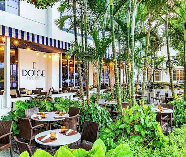 En udendørs restaurant med flere borde og stole omgivet af frodigt grønt, med et skilt for 'Dolce Italian' synligt på bygningen. (AI-genereret tekst)