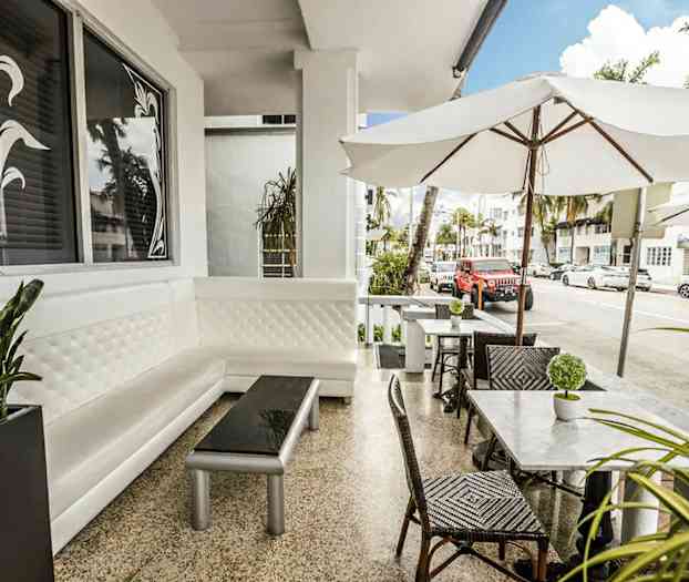 En udendørs servering med hvide møbler og parasoller i et bymiljø. (AI-genereret tekst)