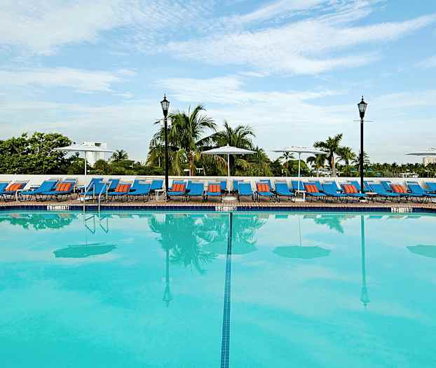 En stor udendørs swimmingpool med liggestole og parasoller omkring, omgivet af palmer. (AI-genereret tekst)