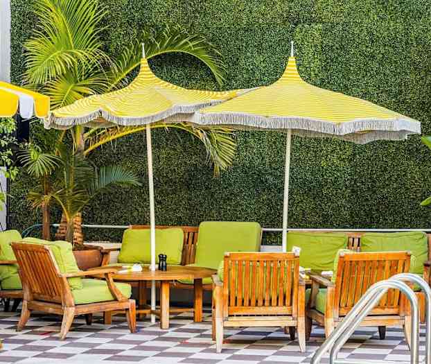 Udendørsstole med grønne puder og træbord under gul- og hvidstribede parasoller, med vegetation i baggrunden. (AI-genereret tekst)