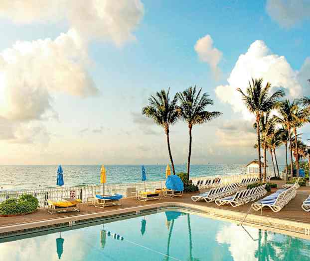 En pool med omkringliggende liggestole og parasoller ved havet, med palmer i baggrunden. (AI-genereret tekst)