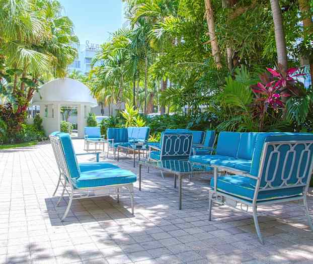 Havemøbler med blå hynder står placeret på en brolagt terrasse, omgivet af palmer og grøn beplantning. (AI-genereret tekst)