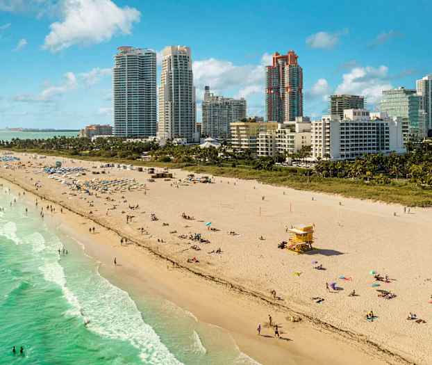 En sandstrand med nogle mennesker, der bader og soler sig tæt på havet. Høje bygninger kan ses i baggrunden. (AI-genereret tekst)