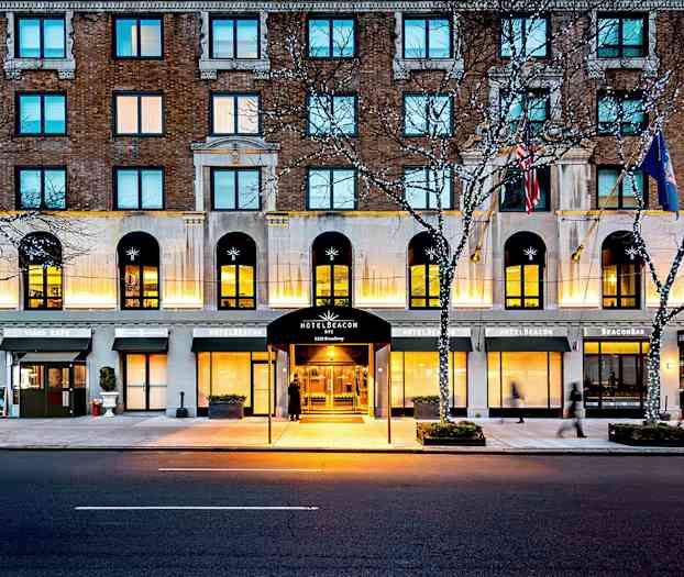 Hotelbygning ved en vej, med indgang markeret som Hotel Beacon, omgivet af cafémiljø og lyskæder i træerne. (AI-genereret tekst)