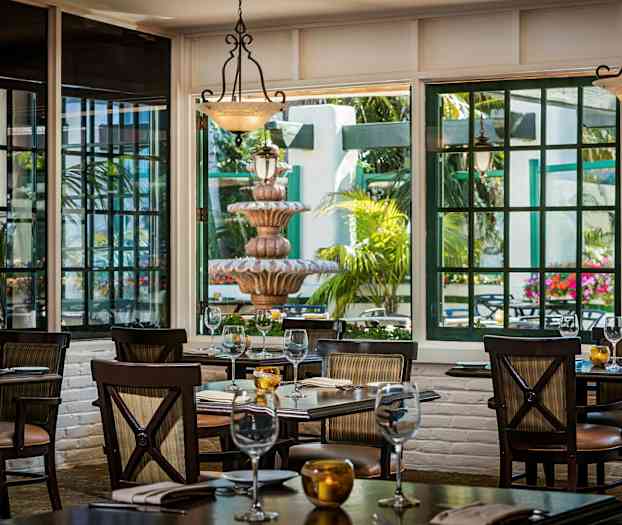 Billede af en restaurant med borde dækket op, store vinduer der slipper lys ind og viser en terrasse med en fontæne og grønne planter. (AI-genereret tekst)