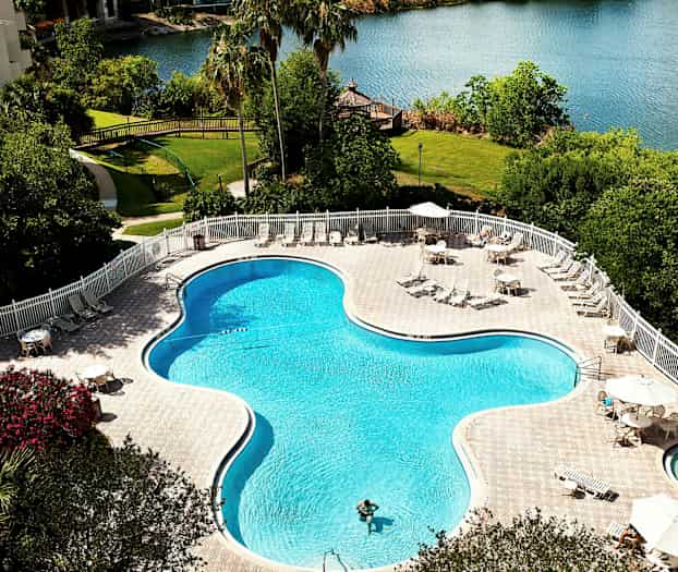 En udendørs swimmingpool omgivet af liggestole og borde med parasoller ved en sø. (AI-genereret tekst)