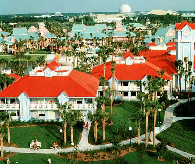 Billede af et feriested med bygninger med rødt tag og lyseblå facader, palmetræer og gangstier. (AI-genereret tekst)