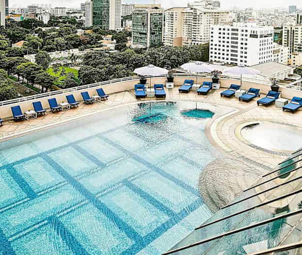 En udendørs pool med blå liggestole og parasoller på en solterrasse, med bylandskab i baggrunden. (AI-genereret tekst)