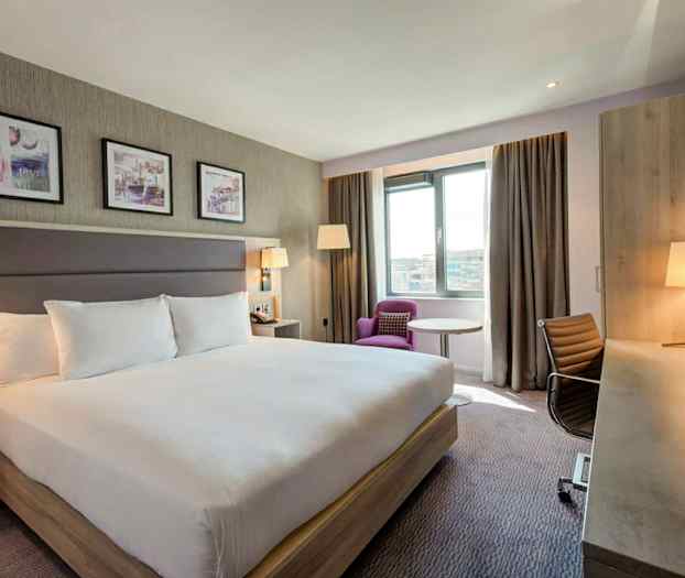 Et hotelværelse med en stor dobbeltseng, skrivebord, stol og en lænestol ved vinduet. (AI-genereret tekst)